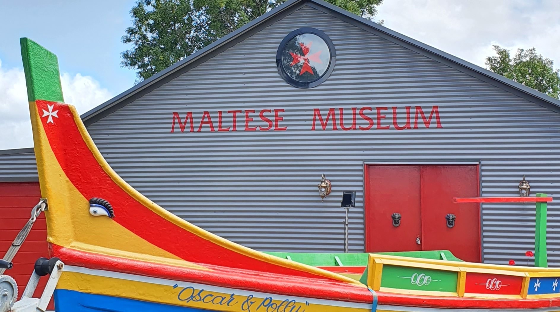 Exterior of the Maltese Museum with a Maltese flag on display. The building is constructed with corrugated iron.
